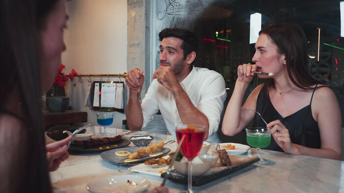 Three people at a table with drinks and food, engaged in a tense conversation causing family drama unintentionally.