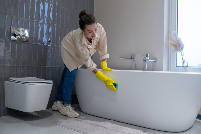 Woman wearing yellow gloves cleaning bathtub in modern bathroom using small tricks to make being a grown-up easier