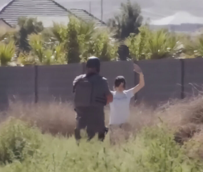 Trans Muslim woman raising hands while facing armed officer outside near a fence in an open grassy area.