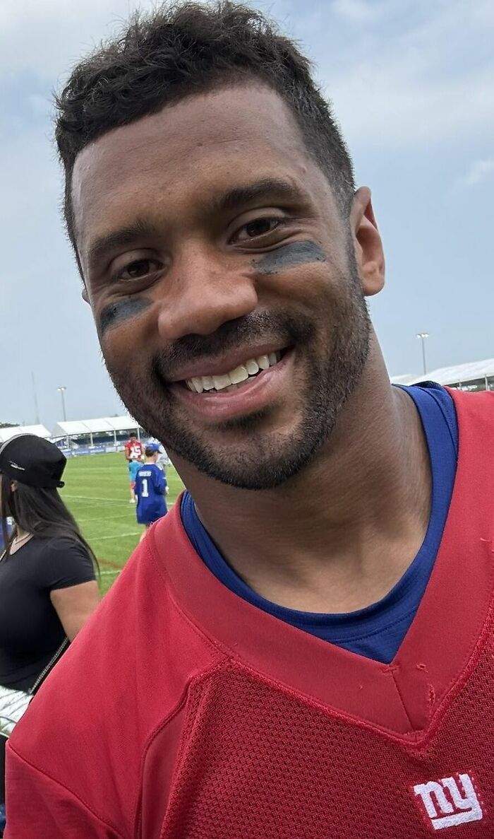 Russell Wilson smiling on a football field wearing a red jersey with eye black during practice session outdoors.
