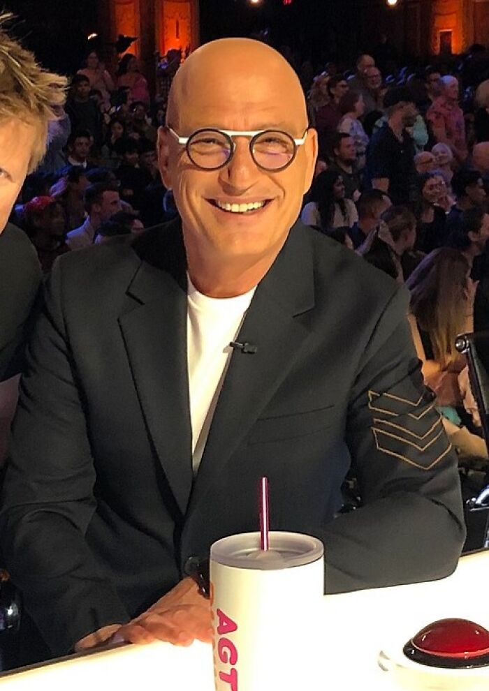 Howie Mandel smiling in glasses and black blazer at a talent show judging panel with audience in background