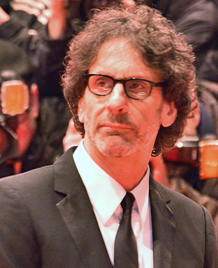 Joel Coen wearing glasses and a black suit at a formal event, highlighting his bio and career achievements.