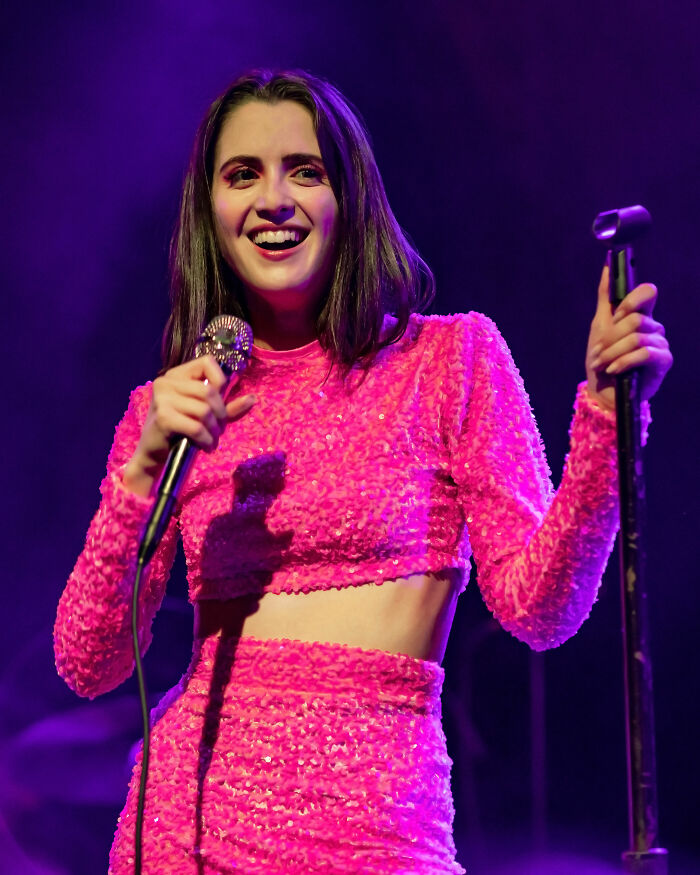 Laura Marano performing on stage in a bright pink outfit, showcasing her bio and career highlights in music.