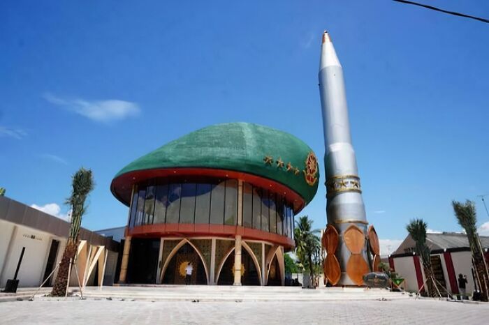 Unique evil building with large green dome and tall rocket-like structure under a clear blue sky in a modern setting.