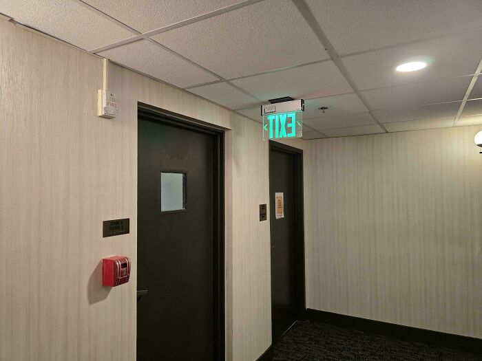 Poorly done job showing an exit sign mounted backward above two black hallway doors in a poorly maintained building.