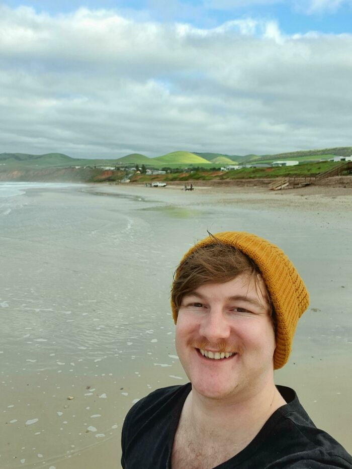 A man wearing a mustard beanie smiling on a beach with green hills in the background, capturing a leap of faith moment.