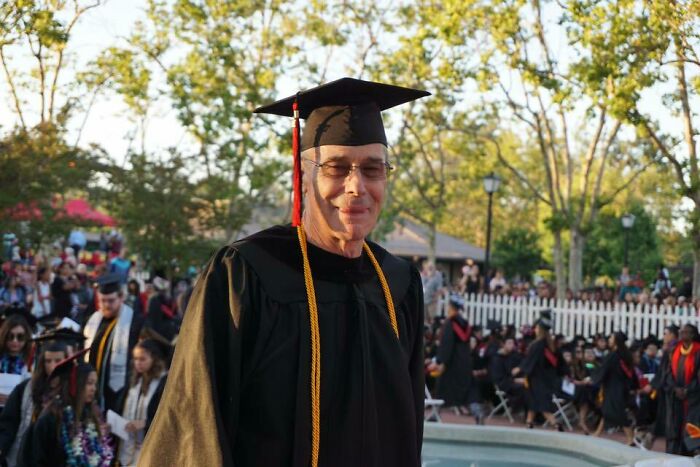 Graduate wearing cap and gown celebrating a leap of faith moment during an outdoor graduation ceremony.