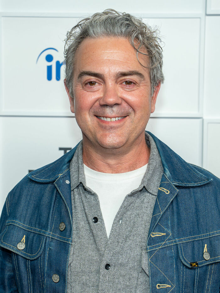 Joe Lo Truglio smiling at an event, wearing a denim jacket and gray shirt against a white background.