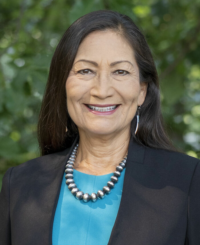 Deb Haaland smiling outdoors, wearing a turquoise top, black blazer, and silver bead necklace in natural light.