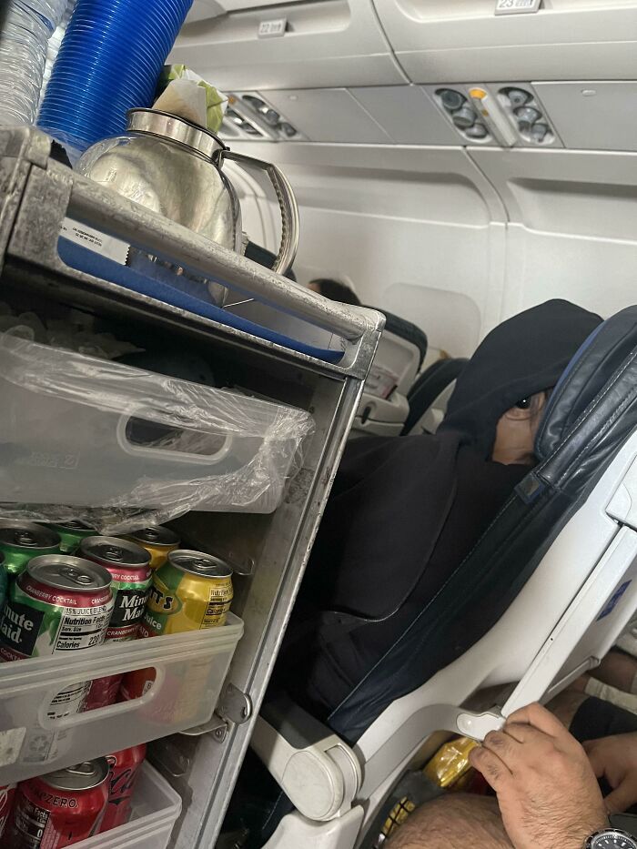 Airplane drink cart with assorted soda cans and a passenger wearing a hood, creating a confusing perspective moment.