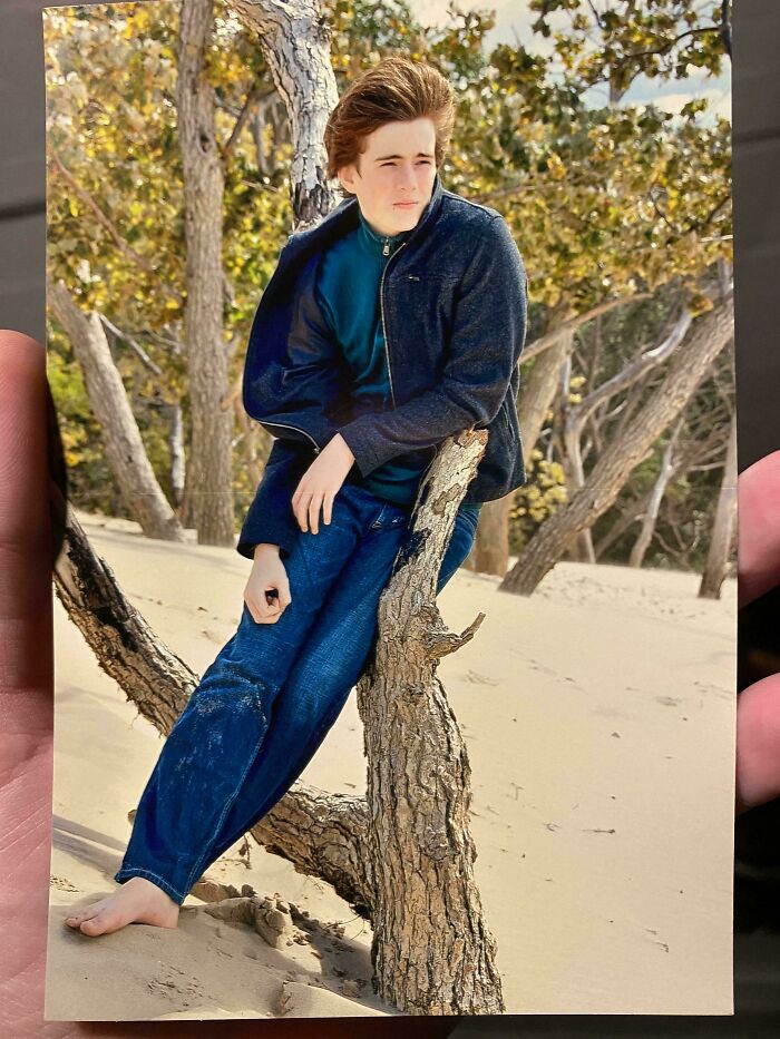 Teen boy in blue jacket and jeans awkwardly leaning on a tree stick in a sandy outdoor setting, childhood photo moment.