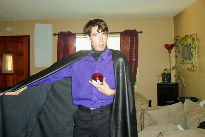 Man in a black cape and purple shirt holding a red ball in a living room, part of hilariously awkward childhood photos.