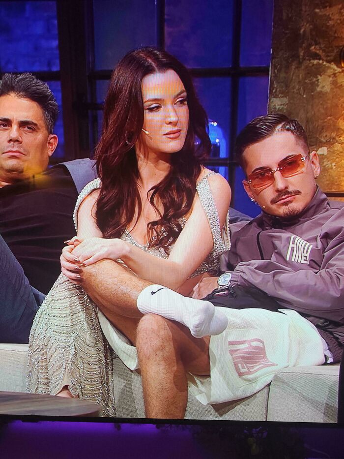 Three people sitting closely on a couch with casual and glamorous outfits in a dimly lit room, creating a confusing visual moment.