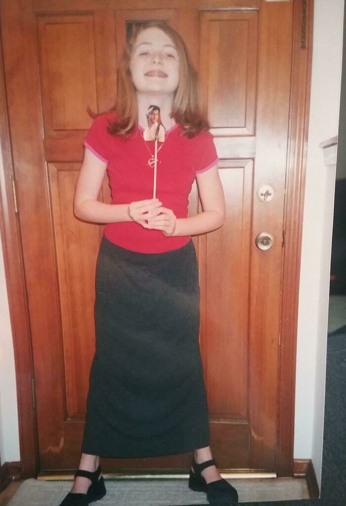 Young girl in a red shirt and long skirt holding a stick with a photo attached, captured in awkward childhood moment.