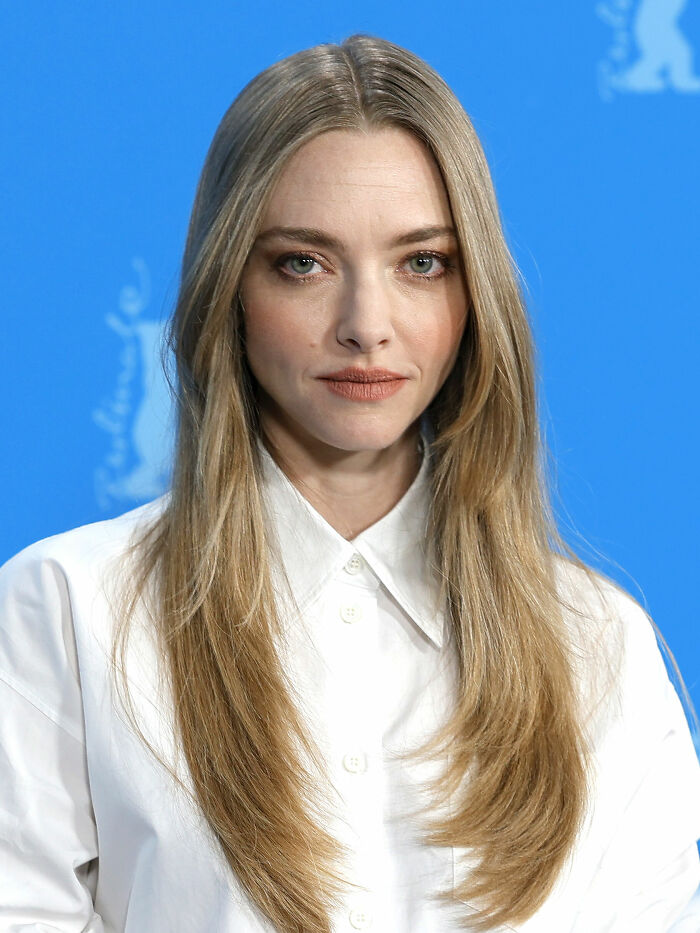 Amanda Seyfried with long blonde hair and white shirt posing against a blue background at an event.