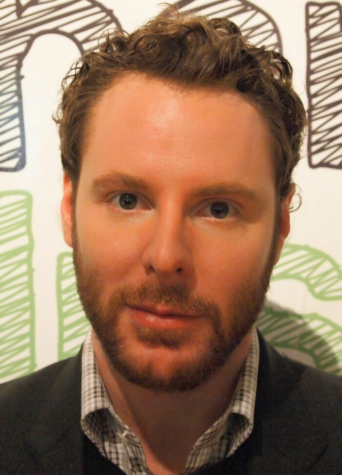 Sean Parker with curly hair and beard, wearing a checked shirt and jacket, posing indoors in front of a patterned background.