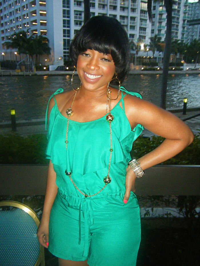 Trina smiling outdoors wearing a green outfit and gold accessories with waterfront and buildings in the background.