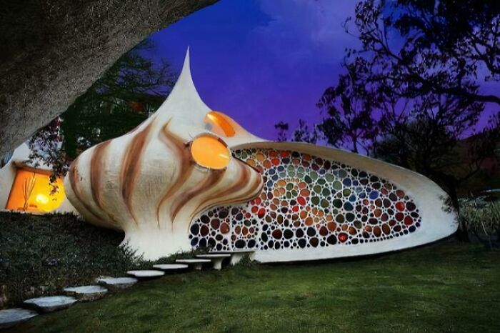 Unique bizarre building shaped like a giant seashell with colorful circular windows at dusk in a natural setting.