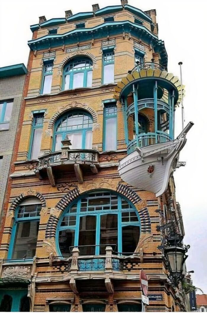 Unique bizarre building with ship-shaped balcony and teal window frames on a vintage brick facade in urban setting.
