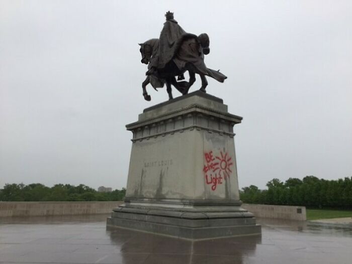 Statue of Saint Louis with red graffiti on its base showing a sun and words, a rage-inducing vandalism image.
