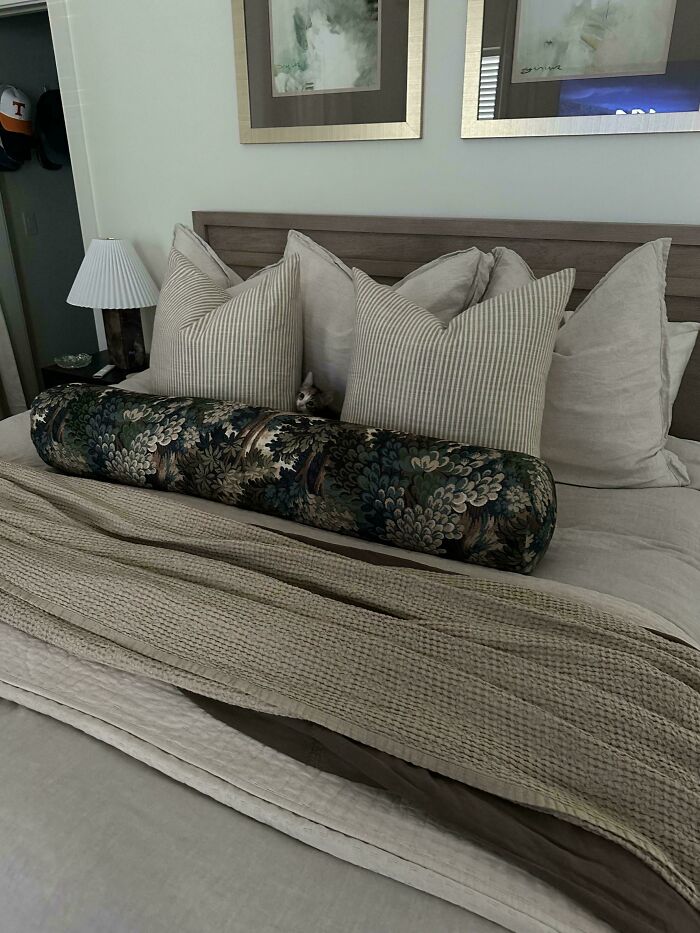 Tiny cat peeking out from behind pillows on a large bed in a cozy bedroom with neutral tones and soft blankets.