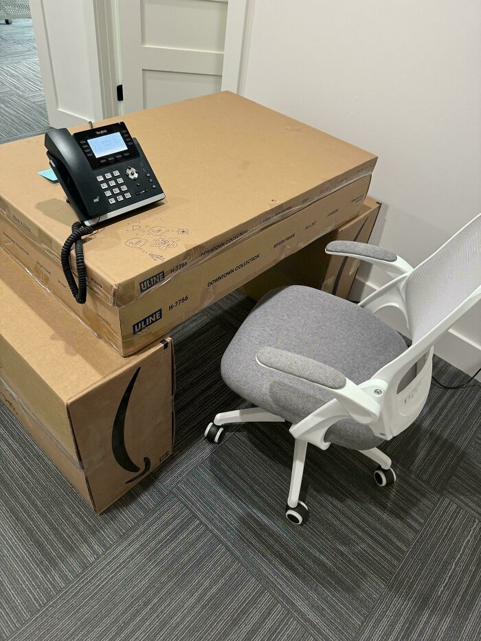 Office makeshift desk made of cardboard boxes with phone on top and gray office chair on carpeted floor.