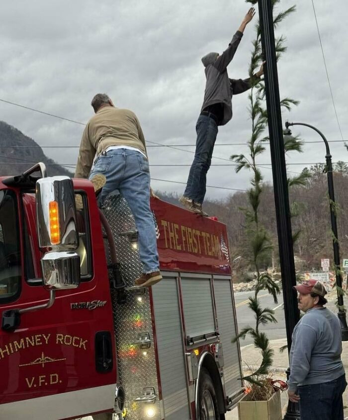 Two men precariously standing on a firetruck attempting to decorate a streetlamp, showcasing poor job performance.