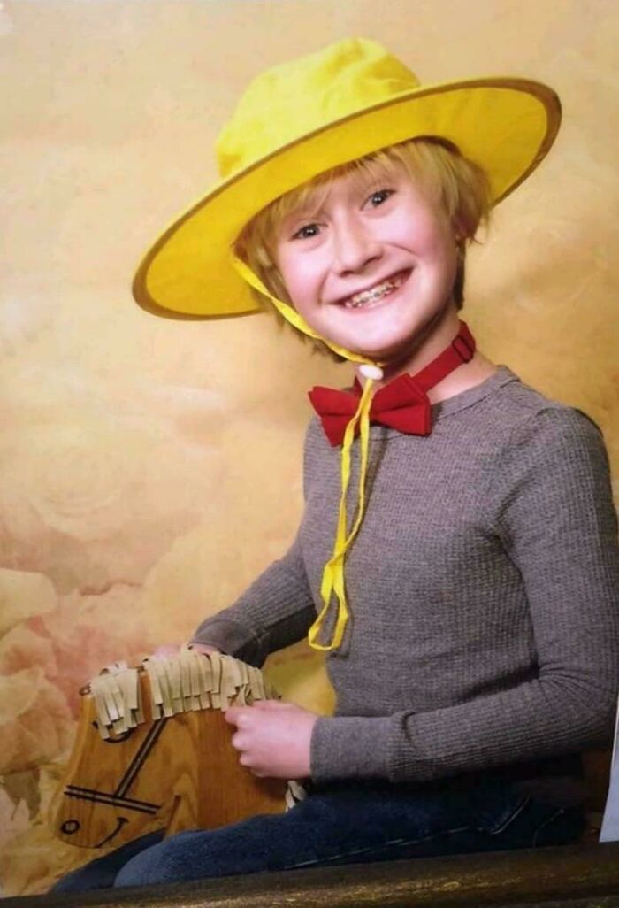 Child wearing a yellow hat and red bow tie, holding a wooden toy horse in a hilariously awkward childhood photo.