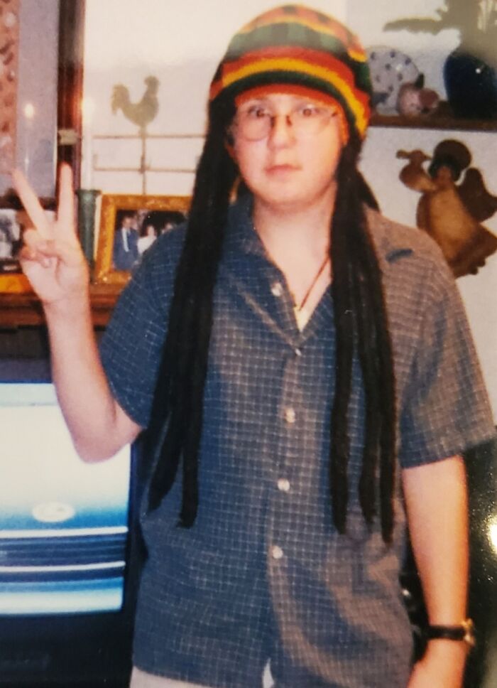 Child wearing a colorful hat with fake dreadlocks and glasses, posing with a peace sign in a humorous childhood photo.