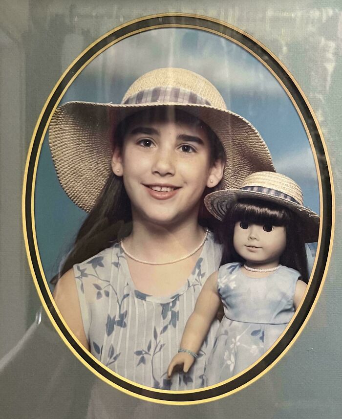 Young girl in a wide-brimmed hat posing with a matching doll in a vintage-style hilariously awkward childhood photo.