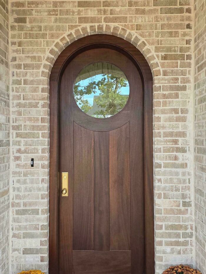 Beautiful wooden front door with round window set in brick wall, showcasing impressive woodworking project craftsmanship.