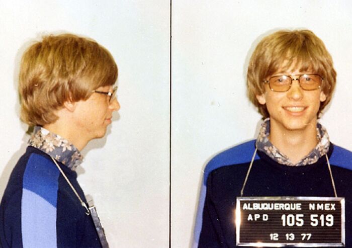 Young man with glasses and patterned shirt in vintage celebrity mugshot, showing arrest details on placard in Albuquerque.