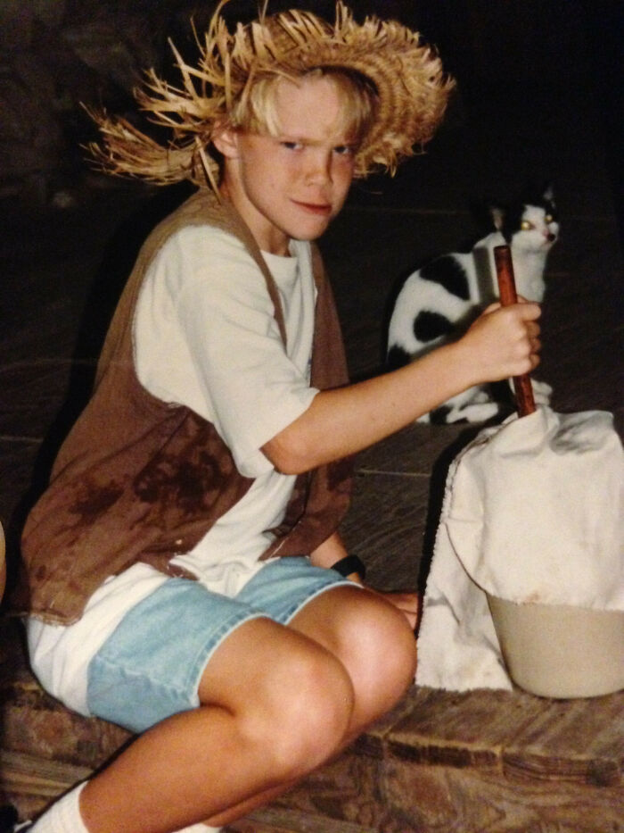 Young boy in a straw hat holding a stick, sitting with a cat, capturing an awkward childhood photo moment.