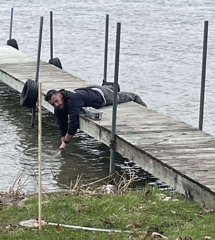 Man lying on a dock reaching into water with his arm blending into the wooden boards, a confusing photo moment.