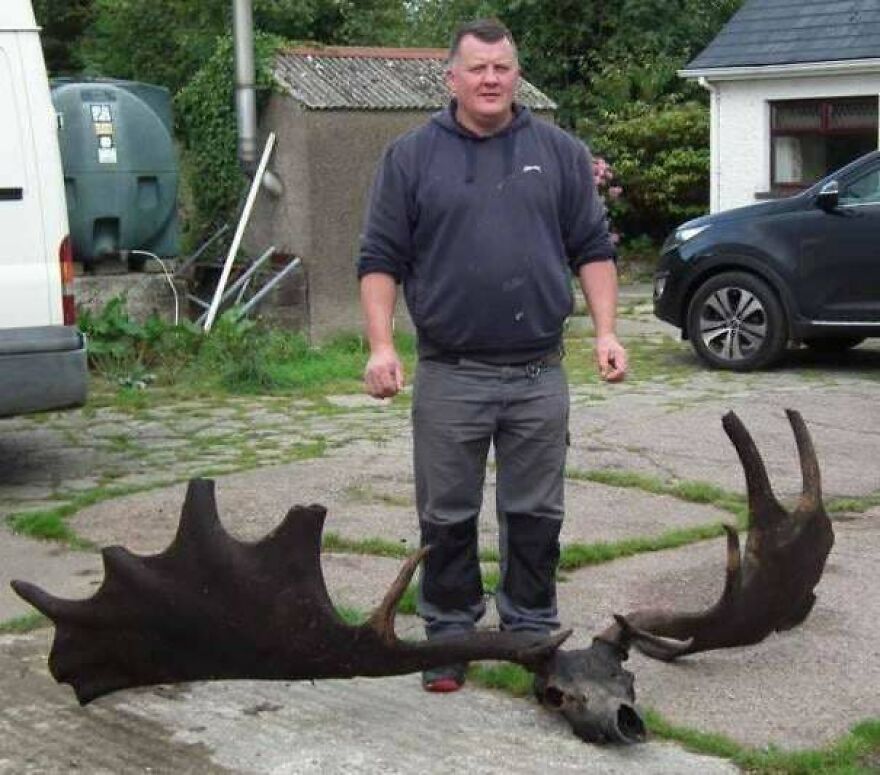 Man standing outdoors next to a gigantic set of antlers, one of the random pictures worth going down a rabbit hole for