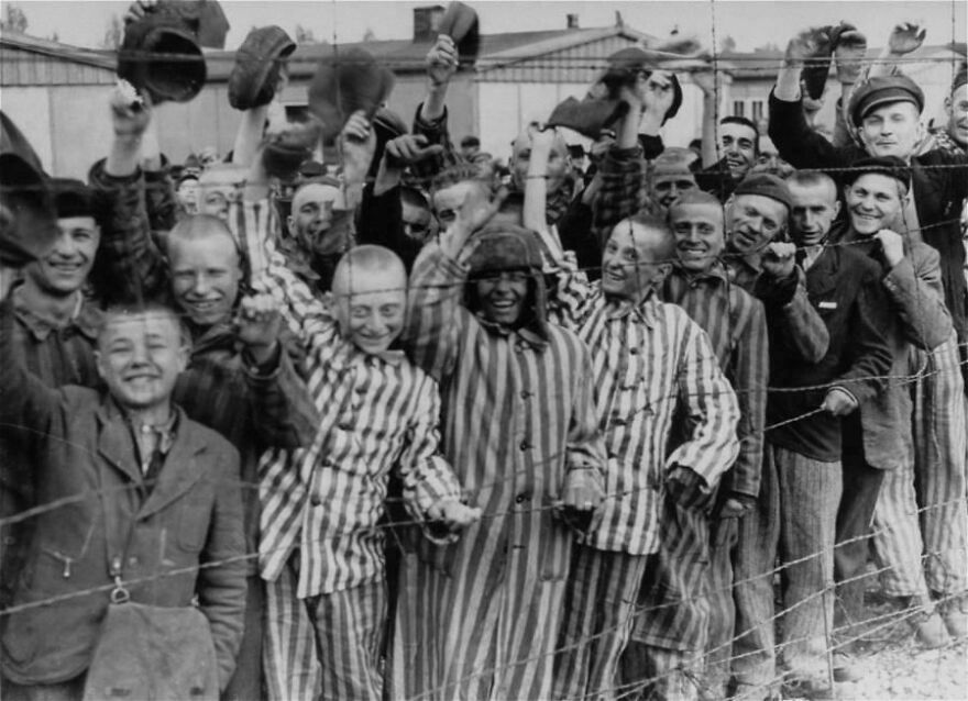 Group of prisoners wearing striped uniforms and hats, smiling and waving behind a barbed wire fence, vintage black and white photo