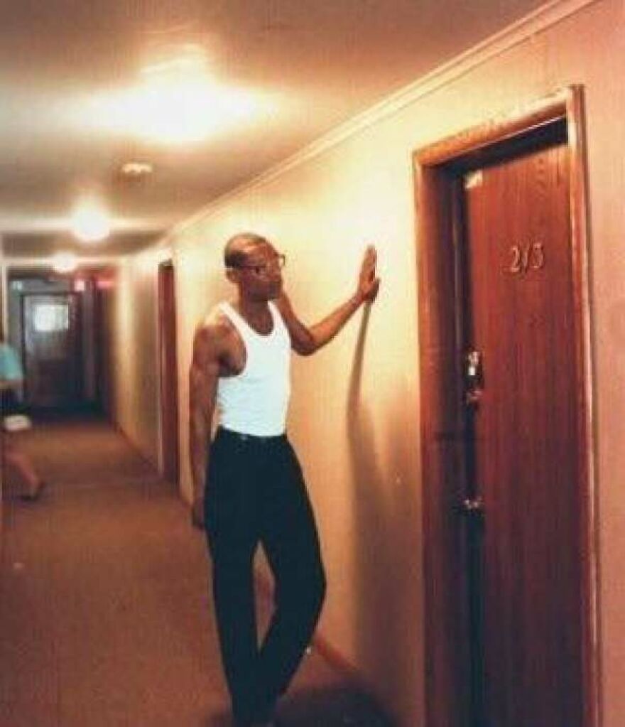 Man in white tank top and black pants standing in a hallway, touching a door numbered 213, a random picture to explore.