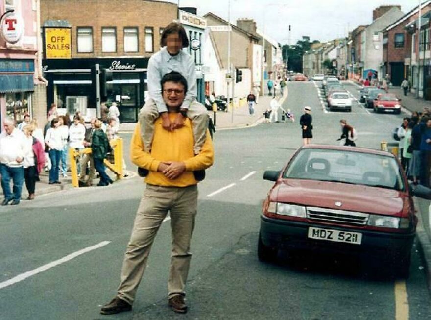 Man in yellow sweater carrying child on shoulders on street with vintage car and crowds, intriguing random pictures to explore