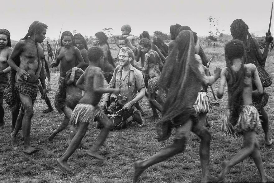Black and white photo of a man with camera surrounded by indigenous people in traditional attire, a captivating random picture.