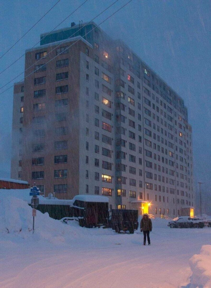 Snow-covered urban scene with a person standing near a lit apartment building, a random picture worth going down a rabbit hole for.