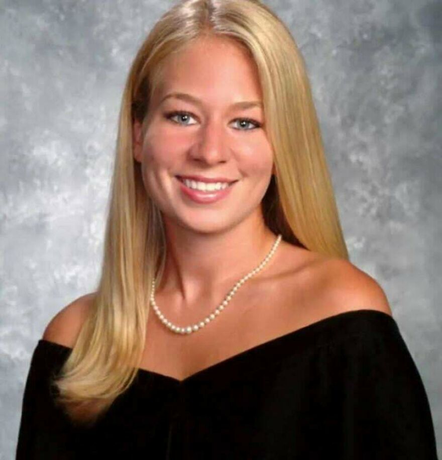 Portrait of a young woman with blonde hair wearing a black off-shoulder top and pearl necklace in a studio setting.
