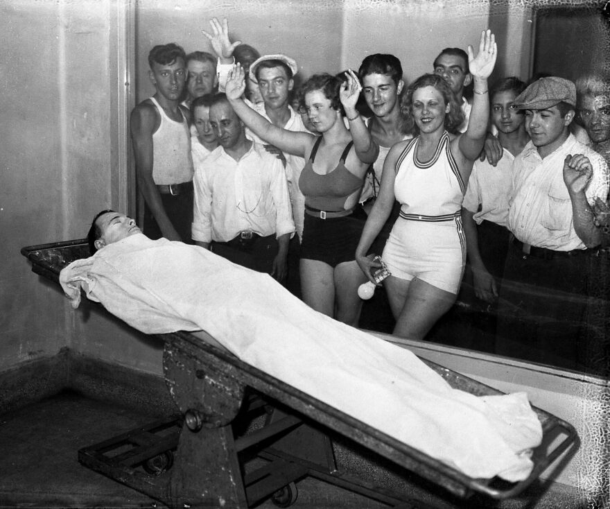 Vintage black and white photo of people observing a woman wrapped in cloth on a tilted table in an unusual scene.