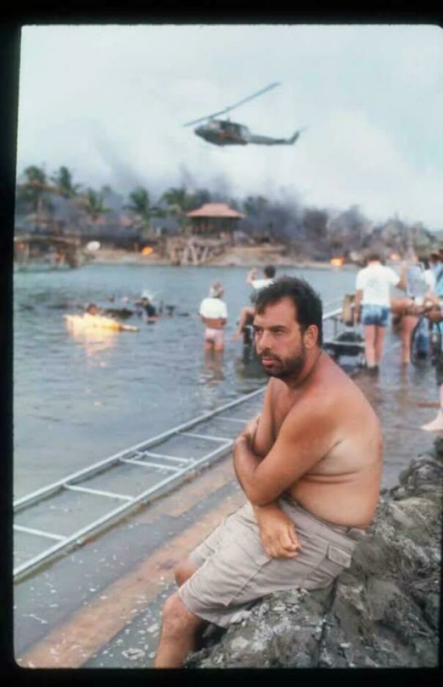 Man sitting shirtless by water with helicopter overhead and people near burning shore in random pictures worth exploring.