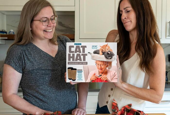 Two women in a kitchen holding a gag gift box labeled Cat Hat, a wearable cat tree, among other gag gifts ideas.