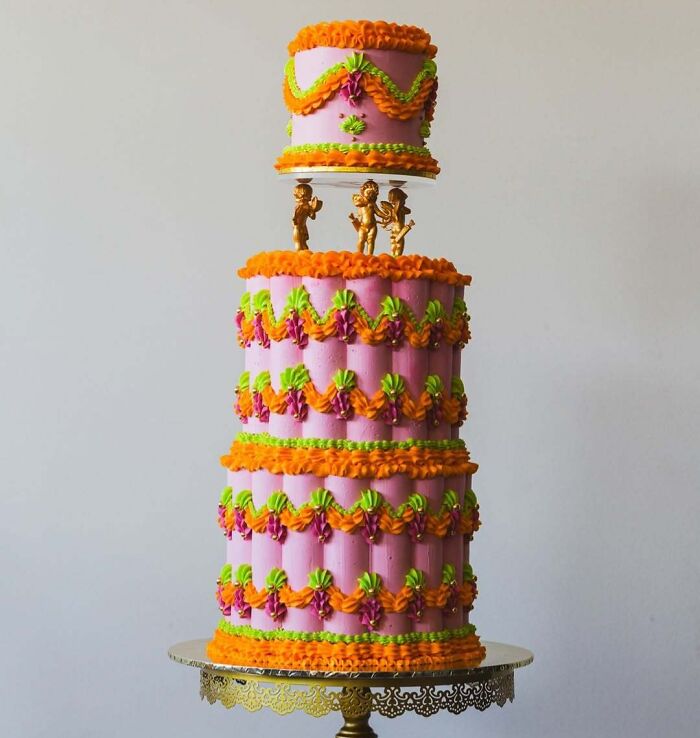 Three-tier masterfully crafted cake decorated with orange, green, and pink frosting and golden figurines on a gold stand.