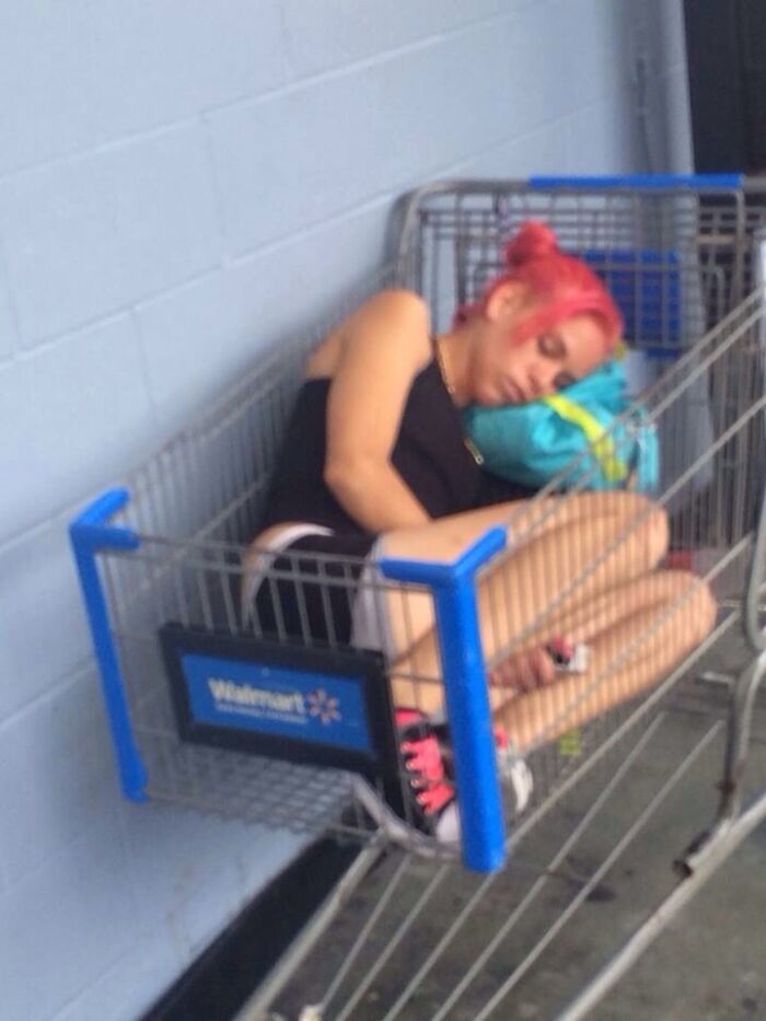 Person with pink hair sleeping curled up inside a Walmart shopping cart outside the store on concrete floor.