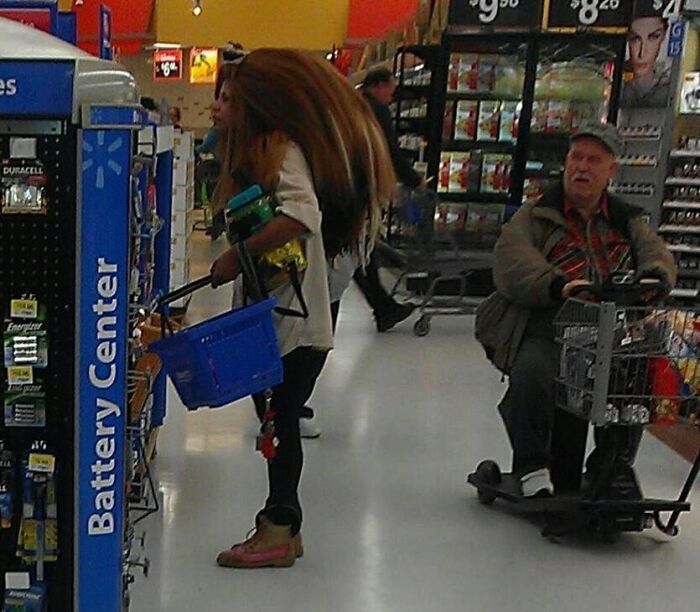 Woman shopping at Walmart battery center while man rides a mobility scooter with a shopping cart in the aisle.
