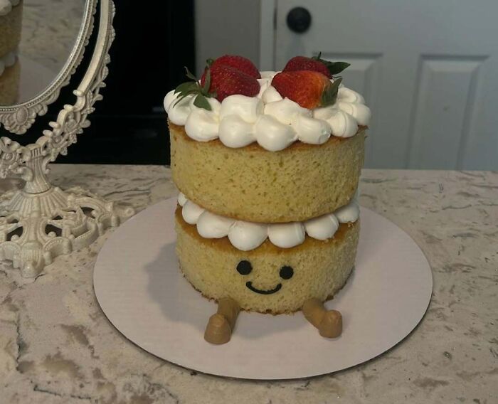 Two-tier masterfully crafted cake with whipped cream, strawberries, and a smiling face decoration on a marble countertop.