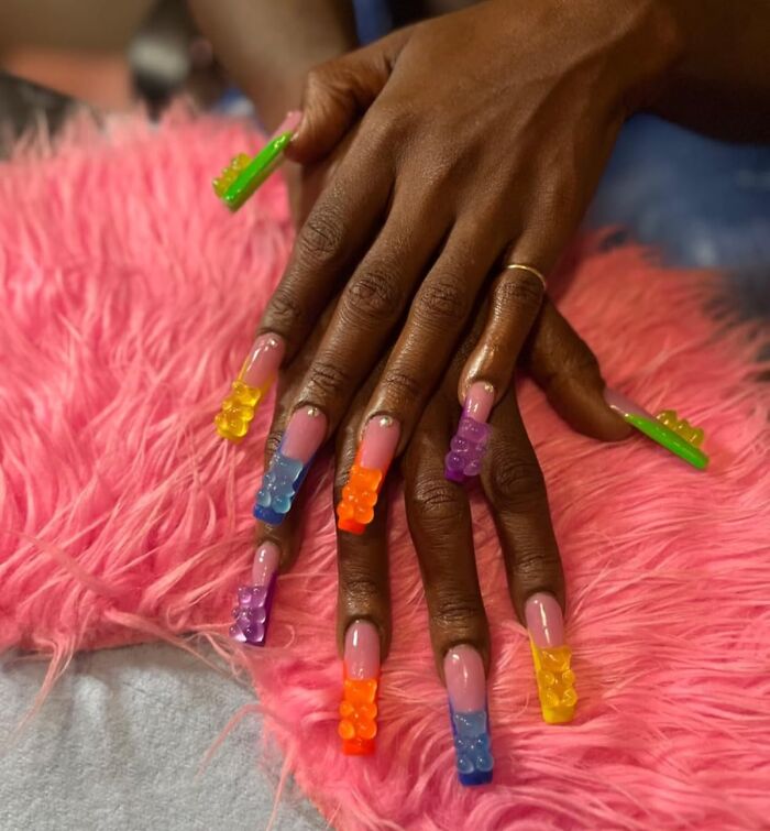 Hands with long nails decorated with colorful gummy bear gag gifts, resting on a pink fuzzy surface.