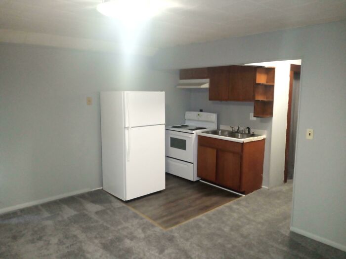 Small kitchen area with white refrigerator, stove, and wooden cabinets in an empty room filled with soft natural light.